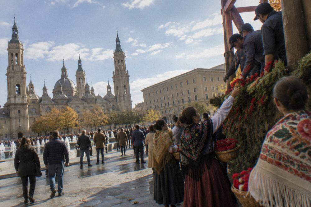 Women in white blouses and colourful shawls handing baskets of red flowers to workers on a tall structure in a crowded square
