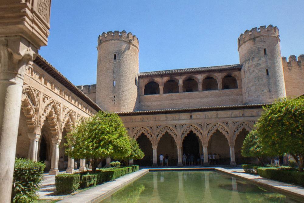 Courtyard of the Aljafería palace with arcades and visitors standing in the shade