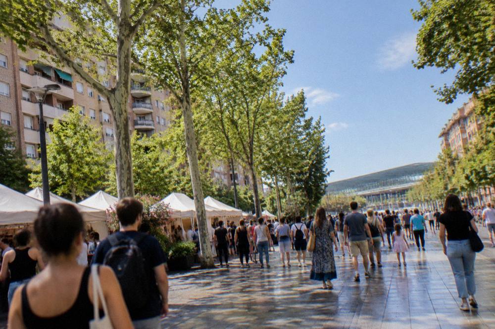 Festival crowds in bright daylight along a tree-lined city promenade with banners overhead