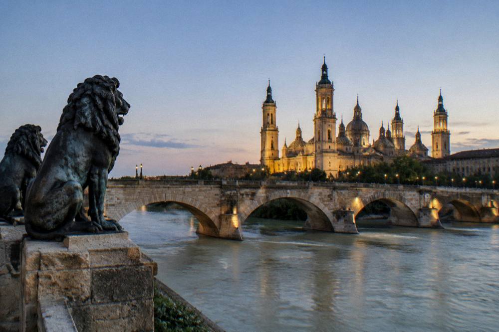 An evening view of a stone bridge over a wide river with a lit basilica tower beyond