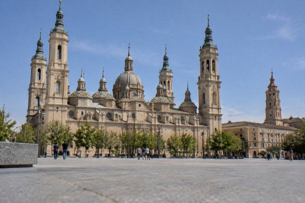 A wide plaza with a large baroque basilica with tiled domes and a second cathedral tower beside it