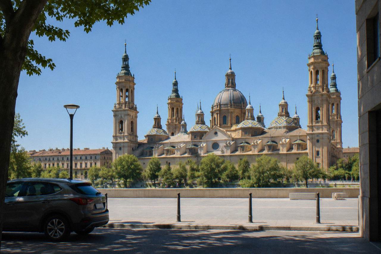 A rental car parked on a quiet street with the Basilica del Pilar rising above the Zaragoza skyline