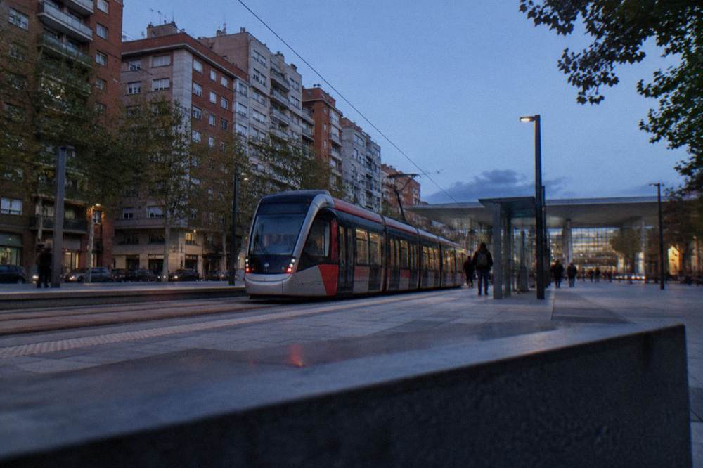 A modern tram waiting at a stop outside a railway station in a Spanish city