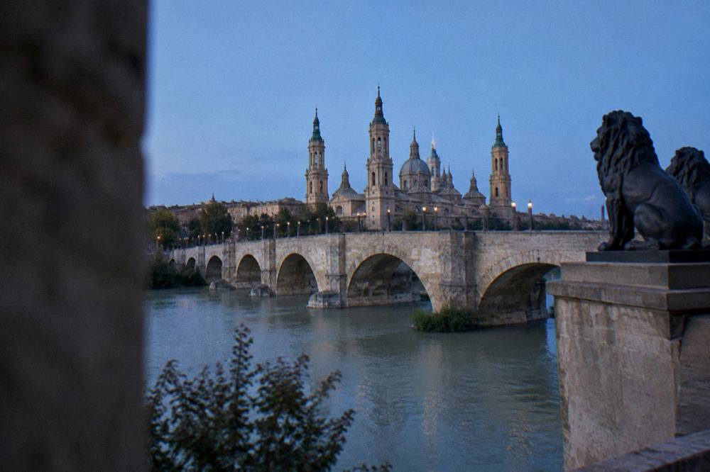 A wide stone bridge over a river with a city skyline of domes and towers behind it