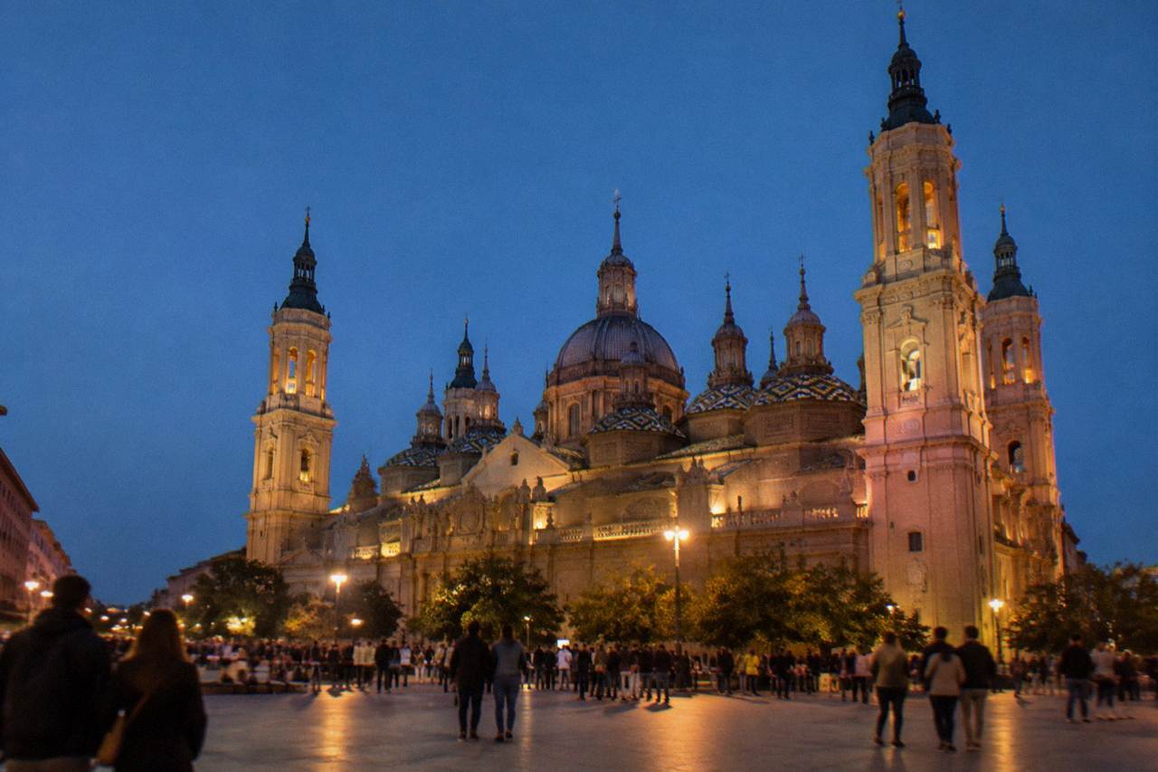 Crowds gathered in a large stone plaza in front of a baroque basilica with tiled domes at dusk