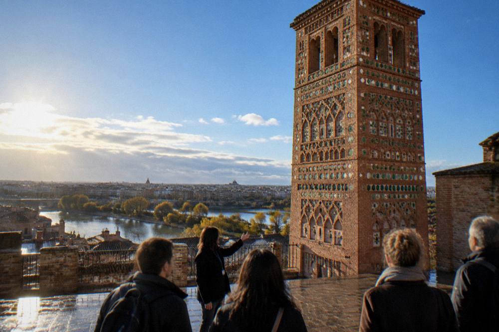A guide speaking to a small group of visitors in a sunlit square beside an ornate brick tower