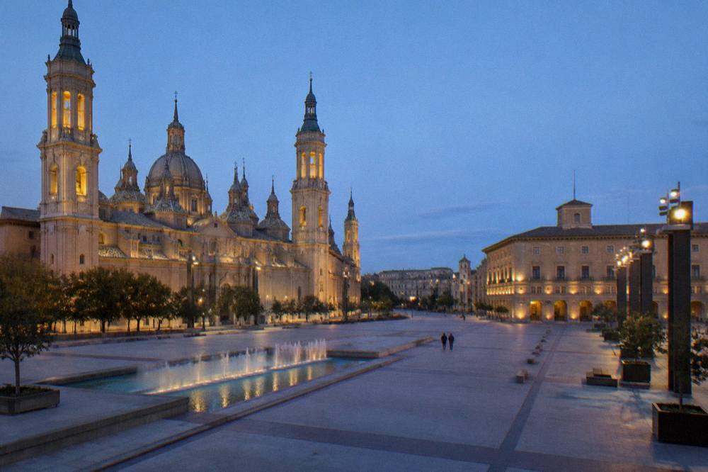 A quiet Zaragoza square at dusk with lit facades and empty benches