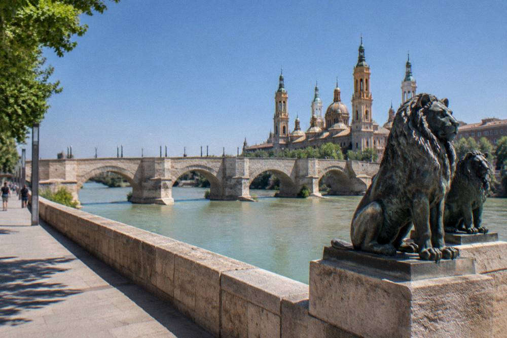 A wide river promenade under a clear blue sky with a historic stone bridge and a basilica dome in the distance