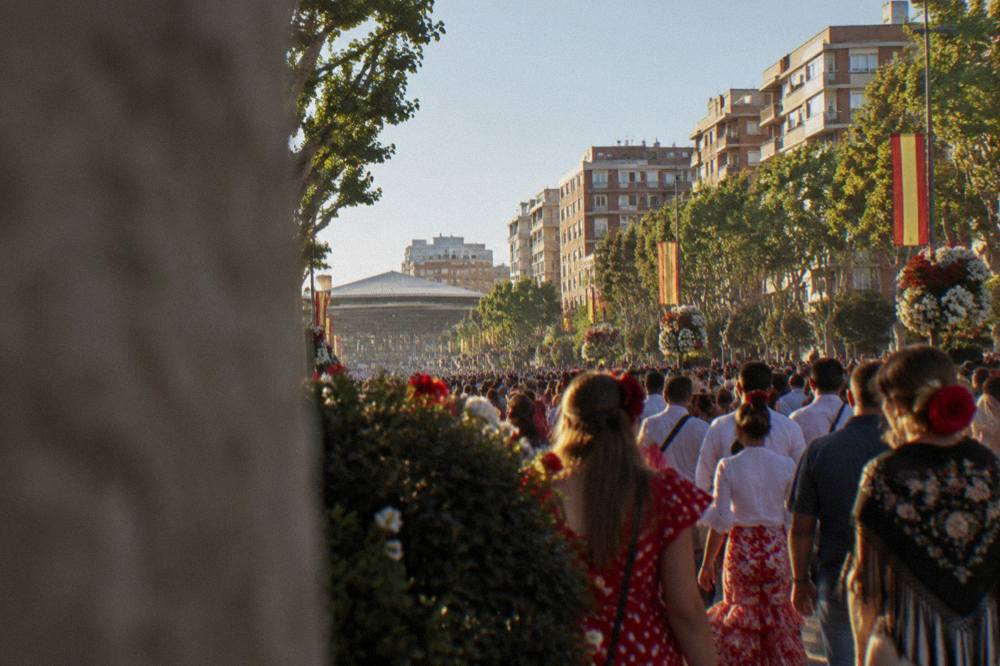 A dense crowd in festive dress filling a broad city avenue lined with flags and flower arrangements