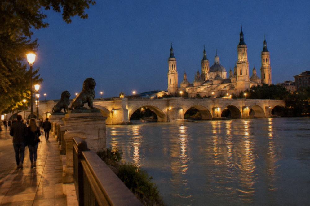 People strolling along the Ebro river promenade at night with lit stone bridges