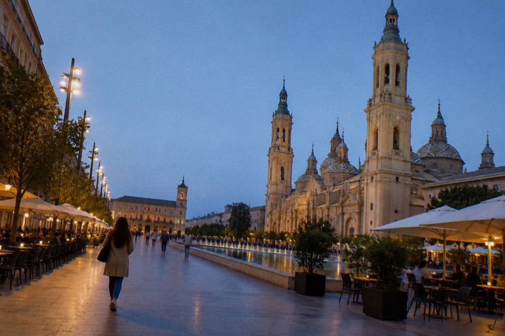 A woman walking alone along a lit evening street in Zaragoza with cafe terraces on both sides