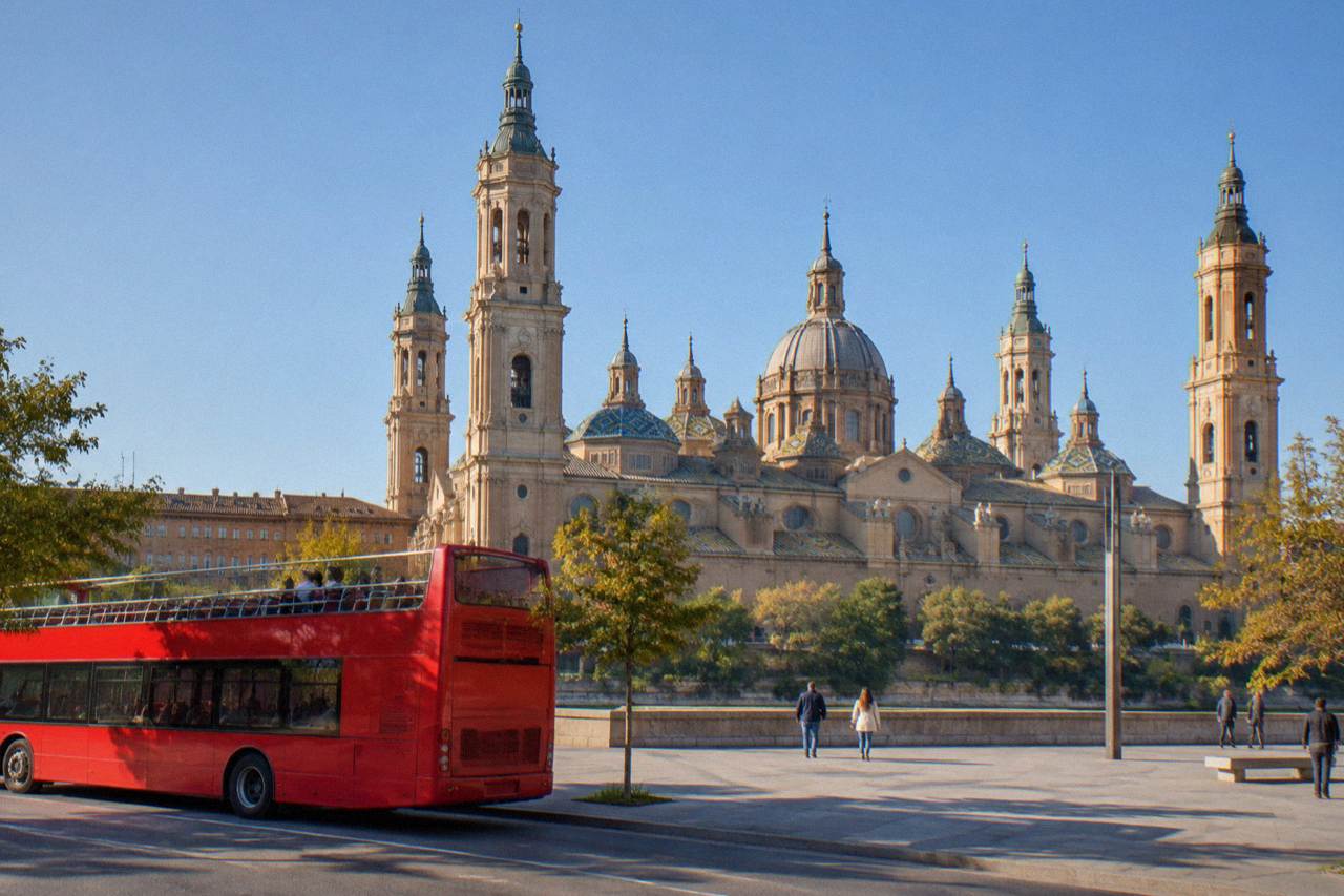 A red open-top tourist bus parked on a sunny street in Zaragoza with the Basilica del Pilar rising behind it
