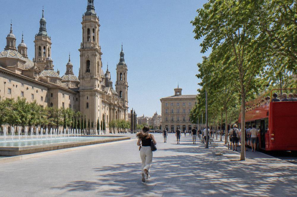 Tourists boarding an open-top sightseeing bus beside a tree-lined avenue in central Zaragoza