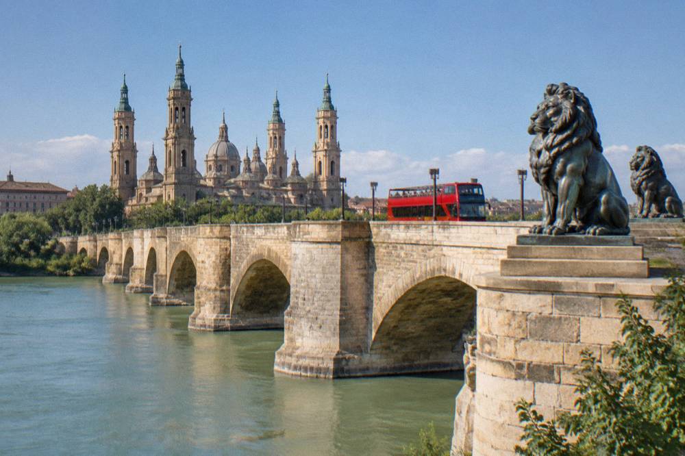 A sightseeing bus driving past the stone arches and lion sculptures of the Puente de Piedra in Zaragoza