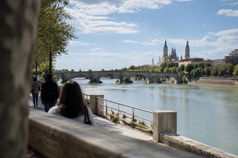 Visitors walking beside the Ebro river in Zaragoza with the stone bridge in view