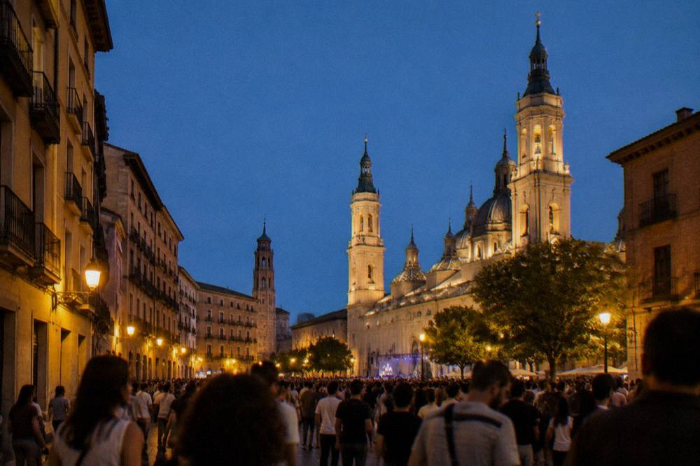 A crowded square at night during a festival with lit church towers above the crowd