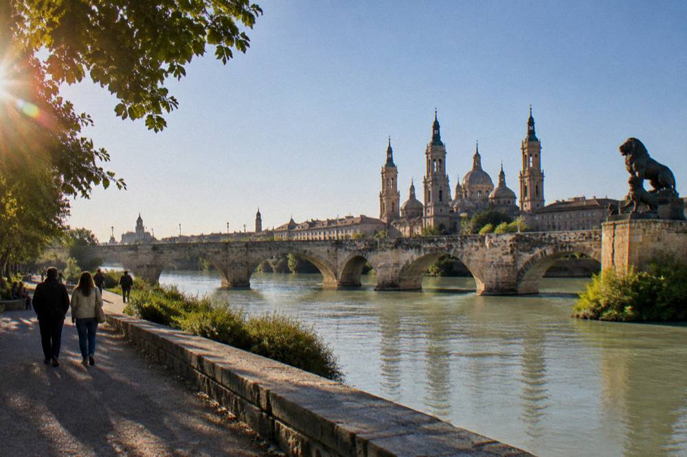 Walkers on the Ebro riverside path with the stone bridge and city skyline along the water
