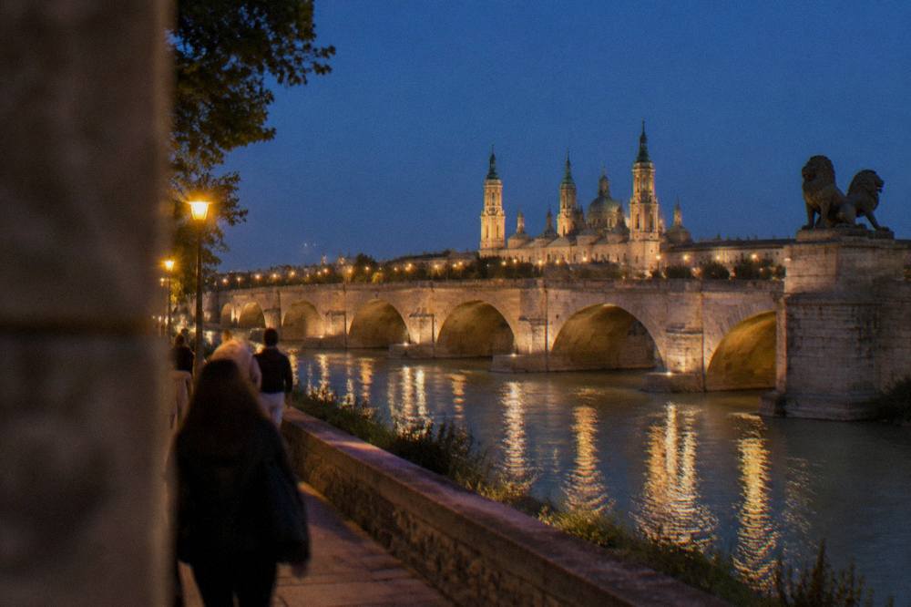 People strolling along the Ebro river path with the stone bridge lit in the background