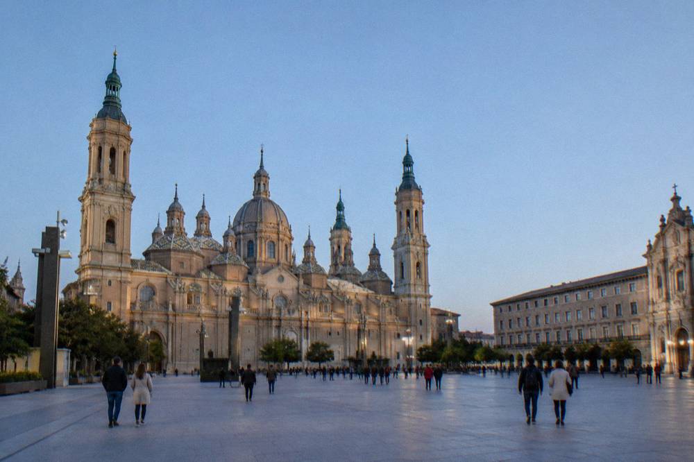 Tourists walking across the wide Plaza del Pilar between the Basilica del Pilar and La Seo cathedral