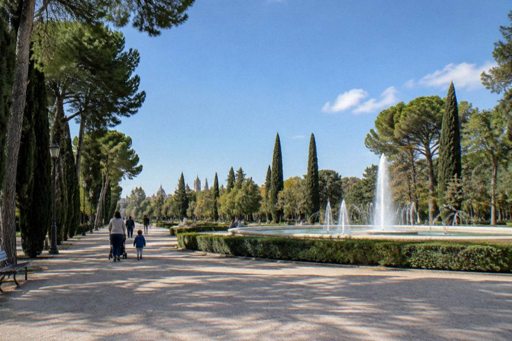 A wide city park with a fountain, tall trees and families walking on gravel paths